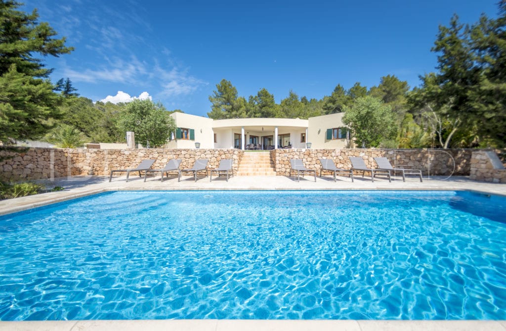 View from the square pool with sun loungers up to the front side of the one floor villa with a round porch