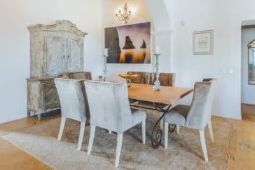 Seating group of a wooden table and velvet chairs on a carpet beside an antique design cupboard and a picture of Ibiza's Es Vedrá