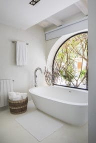 Oval white bathtub beside curved big window with plants