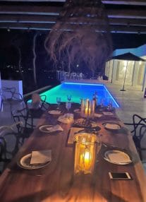 Outdoor dining table with candles in front of Illuminated pool and house at night