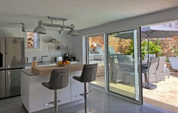Open kitchen with 2 bar stool beside outdoor area with dining table covered by parasol